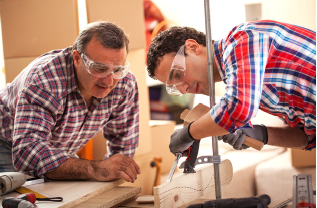 Father carpenter teaches his son how to use a chisel to shapes a wooden plank.