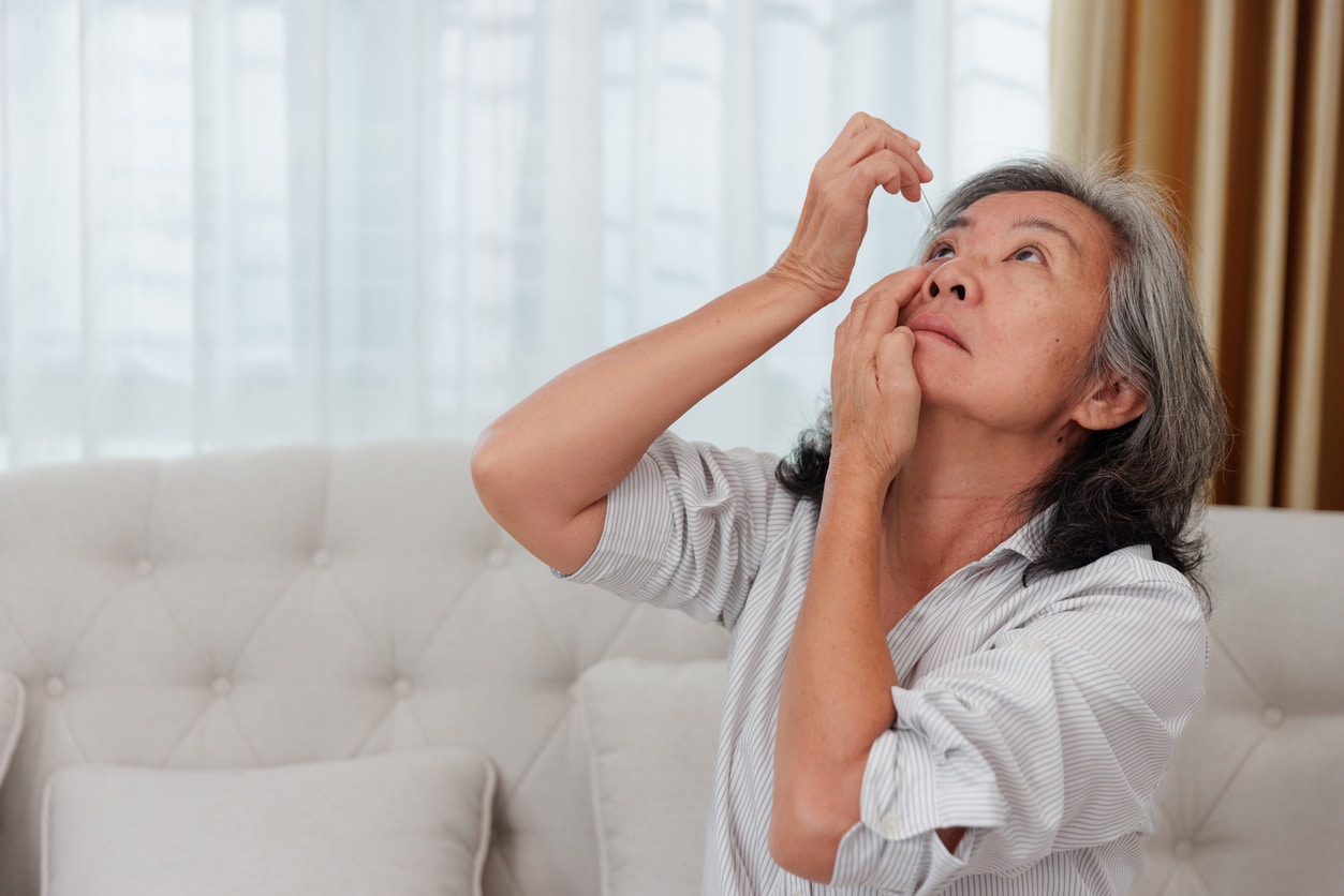 Woman pulling at her eyelid skin Older woman applying prescription eye drops at home for glaucoma management.